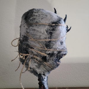 May include: A handcrafted, eerie sculpture of a head wrapped in burlap and twine. The head is painted in shades of gray and black, with small, dark horns. The sculpture has a rough, textured appearance, evoking a sense of mystery and the macabre.