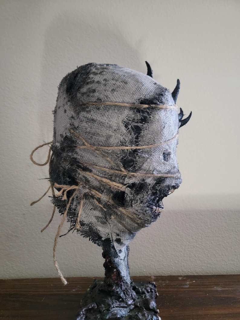 May include: A handcrafted, eerie sculpture of a head wrapped in burlap and twine. The head is painted in shades of gray and black, with small, dark horns. The sculpture has a rough, textured appearance, evoking a sense of mystery and the macabre.