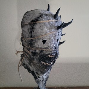 May include: A handmade, textured mask with a burlap-like appearance. The mask features dark, jagged horns and is bound with twine. The mouth area is detailed with a dark, textured material, and the eyes are cut out. The mask is mounted on a dark, textured base.