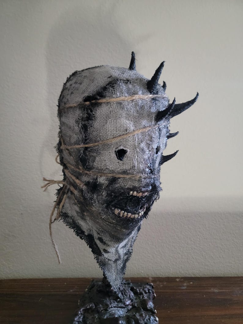 May include: A handmade, textured mask with a burlap-like appearance. The mask features dark, jagged horns and is bound with twine. The mouth area is detailed with a dark, textured material, and the eyes are cut out. The mask is mounted on a dark, textured base.