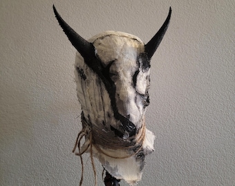 May include: A sculpted head mask featuring a textured off-white fabric covering with distressed black paint markings. The mask has two prominent, curved black horns extending from the top and is wrapped with thin, frayed twine around the neck area.