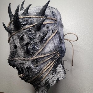 May include: A handmade, textured mask with a distressed appearance. The mask is primarily gray with black accents and features several black, pointed horns. It is wrapped with twine and has a rough, torn texture, suggesting a horror or fantasy theme.