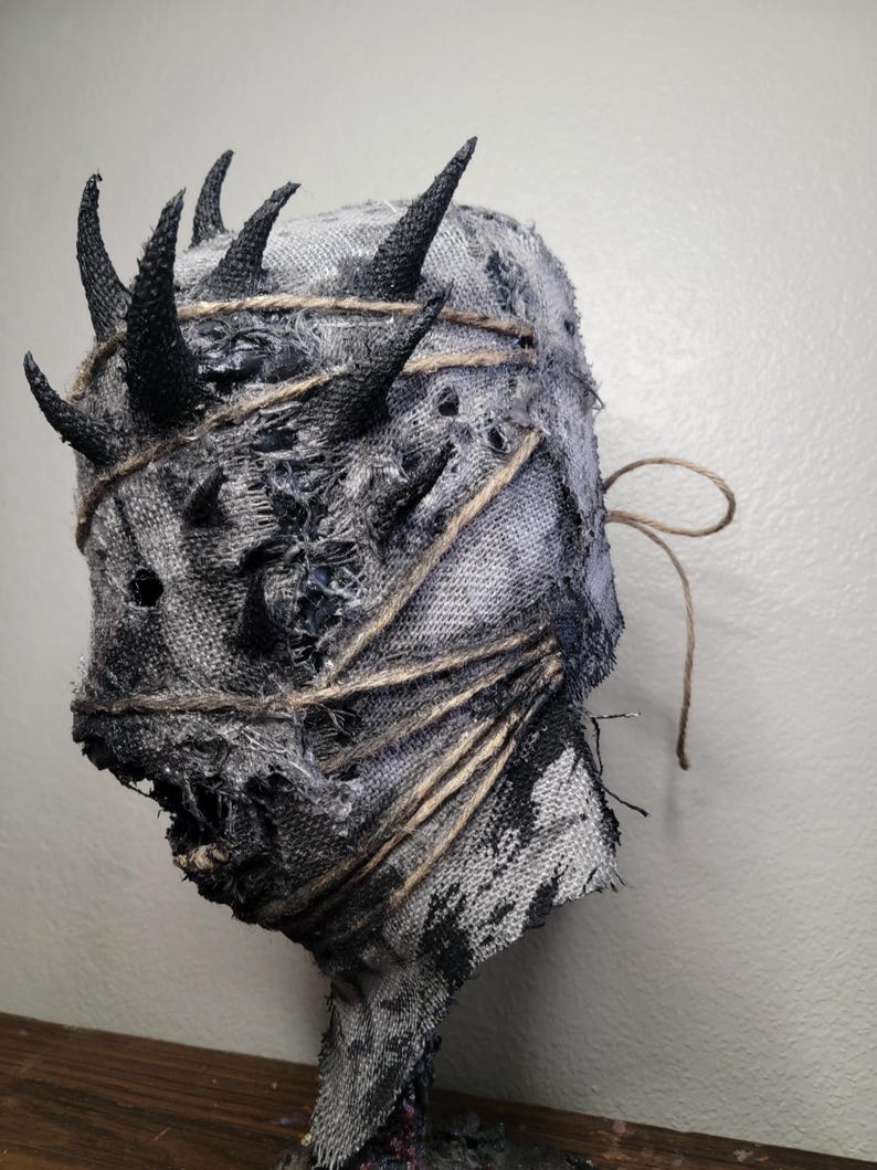 May include: A handmade, textured mask with a distressed appearance. The mask is primarily gray with black accents and features several black, pointed horns. It is wrapped with twine and has a rough, torn texture, suggesting a horror or fantasy theme.