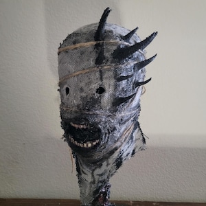 May include: A grotesque mask with a burlap-like texture, featuring black accents and protruding spikes. The mask has a stitched-on appearance, with a wide, toothy grin and dark eye holes. The base is a dark, textured material.