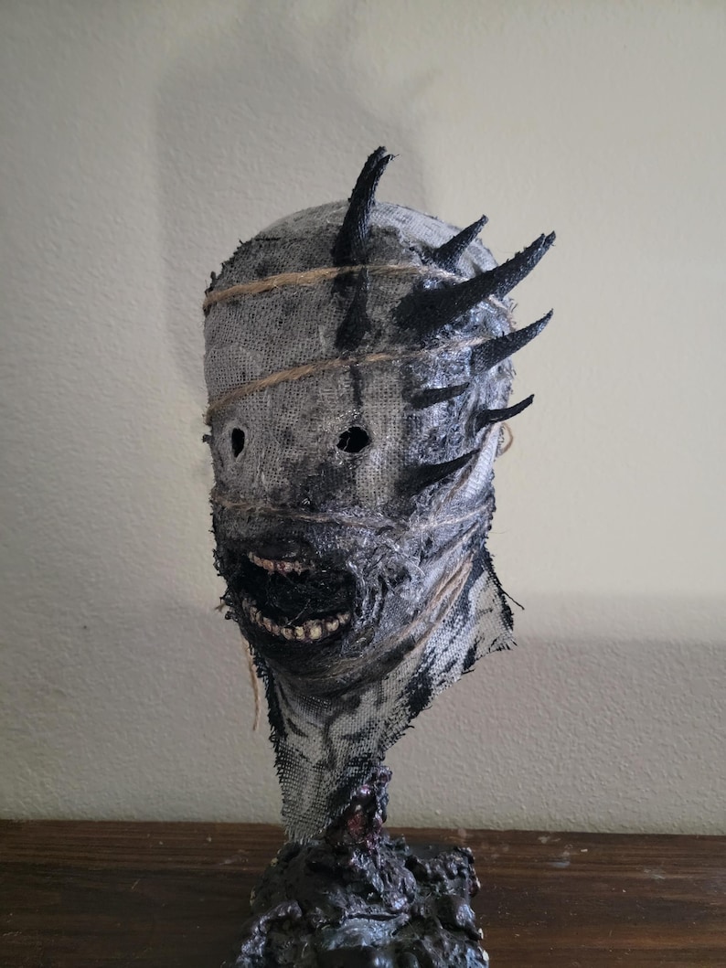 May include: A grotesque mask with a burlap-like texture, featuring black accents and protruding spikes. The mask has a stitched-on appearance, with a wide, toothy grin and dark eye holes. The base is a dark, textured material.