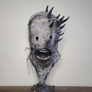 May include: A grotesque, handmade mask with a burlap texture, featuring jagged black spikes and a stitched-up appearance. The mask has a wide, menacing grin and is bound with twine. It rests on a dark, textured base, creating a chilling, horror-themed aesthetic.
