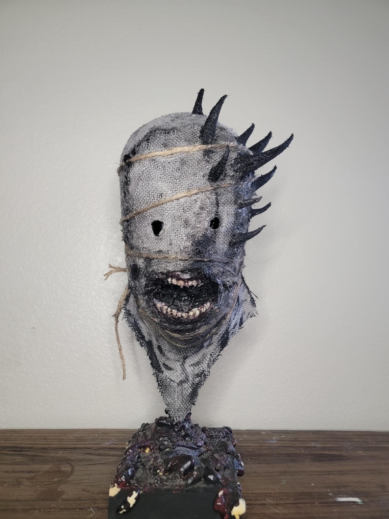 May include: A grotesque, handmade mask with a burlap texture, featuring jagged black spikes and a stitched-up appearance. The mask has a wide, menacing grin and is bound with twine. It rests on a dark, textured base, creating a chilling, horror-themed aesthetic.