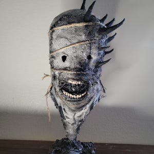 May include: A handcrafted, textured mask with a menacing expression. The mask features a burlap-like material, dark accents, and protruding spikes. The mouth is open, revealing teeth. The mask is mounted on a black base, creating a spooky, Halloween-themed decoration.