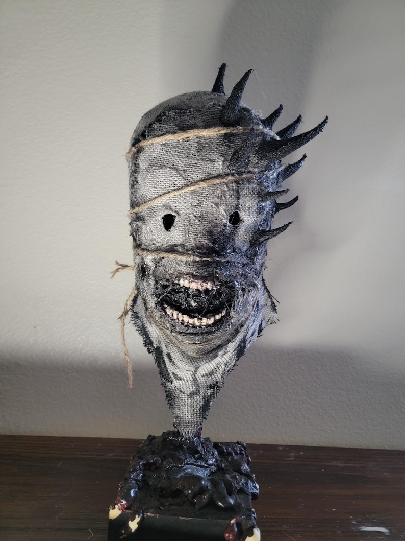 May include: A handcrafted, textured mask with a menacing expression. The mask features a burlap-like material, dark accents, and protruding spikes. The mouth is open, revealing teeth. The mask is mounted on a black base, creating a spooky, Halloween-themed decoration.