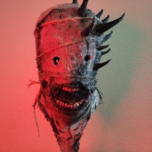 May include: A burlap mask with a menacing design. The mask features a stitched burlap texture, dark eye holes, and a wide, toothy grin. Black spikes protrude from the top. The mask is tied with twine and has a dark, textured base.
