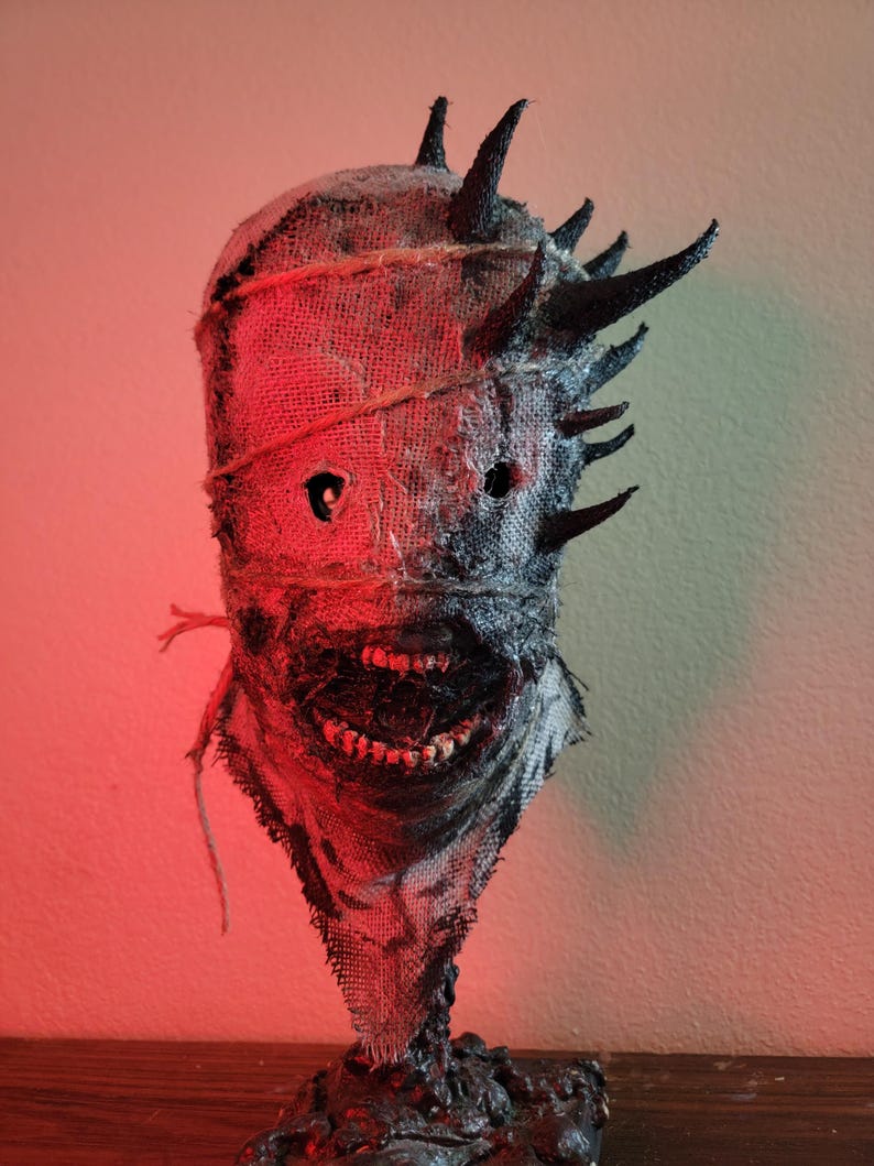 May include: A burlap mask with a menacing design. The mask features a stitched burlap texture, dark eye holes, and a wide, toothy grin. Black spikes protrude from the top. The mask is tied with twine and has a dark, textured base.