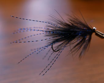 May include: Close-up of a black fishing fly with a curved hook and a cluster of black feathers. The fly has delicate blue accents and is held by a silver tool, showcasing its intricate design. The background is a blurred brown.