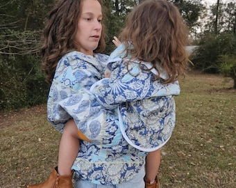 May include: A woman wearing a blue and white paisley print shirt is holding a young girl in a matching shirt. The woman is wearing brown boots and blue jeans. The girl is wearing brown shoes.