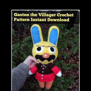 May include: Crochet pattern for Gaston the villager from Animal Crossing. The pattern is for a yellow bunny with blue ears, a brown and white shirt, and a red skirt. The pattern is available for instant download.