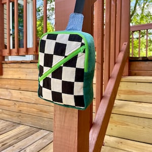 May include: A crossbody sling bag featuring a black and white checkered front panel with a diagonal lime green zipper. The bag has solid dark green side panels, a blue denim-textured top accent, and a black adjustable nylon shoulder strap.