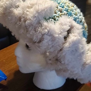 May include: A white faux fur hat with a crocheted blue, green, and yellow brim. The hat is on a white mannequin head.