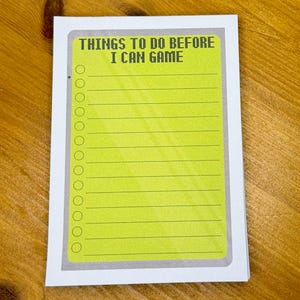 Op de afbeelding: Notitieblok met een felgele pagina en de tekst "THINGS TO DO BEFORE I CAN GAME" in een pixelachtige lettertype. Het notitieblok heeft een grijze rand en een reeks cirkels aan de linkerkant, met horizontale lijnen om op te schrijven.