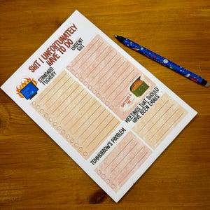 May include: A white notepad with a humorous design and a blue pen. The notepad has sections labeled "Standard Fuckery," "Urgent Shit," and "Meetings That Should Have Been Emails." The pen has a blue barrel with a space-themed design.