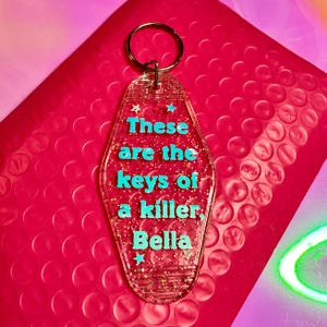Puede incluir: Llavero acrílico transparente con forma de llave de motel, con el texto "These are the keys of a killer Bella" en turquesa. Tiene un llavero plateado y detalles de purpurina. Fondo rosa.