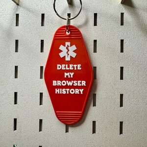 May include: A red keychain with the text "DELETE MY BROWSER HISTORY" in white. The keychain has a white medical symbol and a silver key ring. The background is a white pegboard.