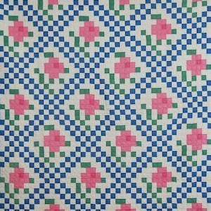 May include: A quilt featuring a repeating pattern of pink pixelated flowers with green stems and leaves, set against a background of small, alternating blue and white squares. The textile displays a grid-like arrangement of floral motifs.