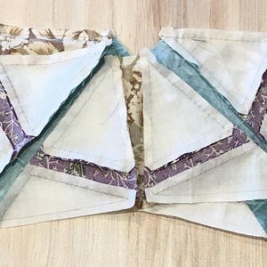 Op de afbeelding: Twee onafgewerkte quiltblokken met een witte achtergrond en verschillende stofresten in tinten paars, teal en bruin. De blokken zijn aan elkaar genaaid en hebben een stervormig ontwerp.