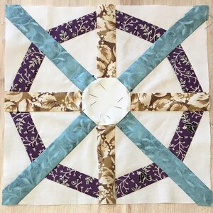 Op de afbeelding: Een quiltblok met een zeshoekig patroon in het midden. Het blok is gemaakt van verschillende stoffen in tinten bruin, paars en blauw. Het midden van het blok is een witte cirkel met een pin erin.