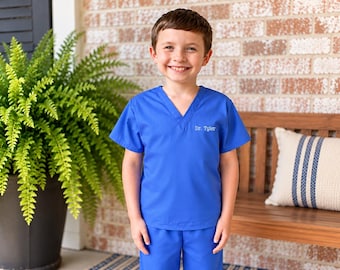 Puede incluir: Un conjunto de casaca con cuello en V y pantalón a juego en color azul brillante. La parte superior tiene mangas cortas y el texto blanco bordado "Dr. Tyler" en el área del pecho izquierdo. La tela es de un color azul sólido y vibrante.