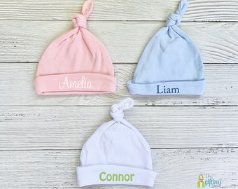 Könnte beinhalten: Drei personalisierte Baby-Mützen in Rosa, Blau und Weiß mit den Namen Amelia, Liam und Connor bestickt.