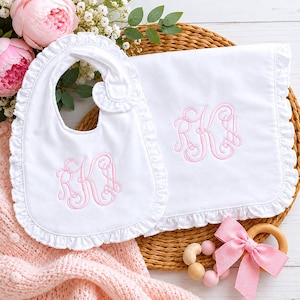 May include: White baby bib and blanket set with pink embroidered initials and ruffled edges. A wooden teething ring with pink beads and a bow is also present. The set is on a woven mat with pink flowers.