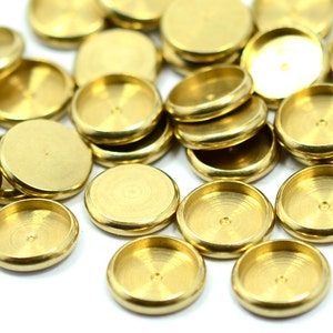 Könnte beinhalten: Eine Sammlung kleiner, runder Metallkomponenten in Messingfarbe. Jede Komponente hat eine flache Oberseite und eine flache, konkave Basis. Die Komponenten sind auf einer weißen Oberfläche verstreut, wobei sich einige überlappen.