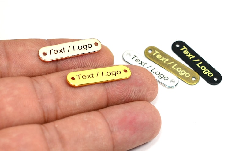 K&ouml;nnte beinhalten: Vier Metall-Etiketten mit abgerundeten Kanten und zwei L&ouml;chern. Die Etiketten sind in Gold, Silber, Messing und Schwarz. Jedes Etikett tr&auml;gt die Gravur "Text / Logo".