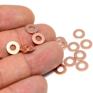 Puede incluir: Un grupo de arandelas de metal de color cobre, algunas están en una mano, otras están esparcidas sobre una superficie blanca.