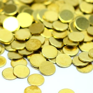 Peut inclure: Gros plan sur une pile de petits disques ronds en métal doré. Les disques sont dispersés sur une surface blanche, créant un motif texturé et visuellement attrayant. Les disques semblent être en laiton ou en alliage similaire.