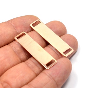 May include: Two rose gold metal bar tags with a rectangular shape and a small hole at each end. The tags are lying on a hand.