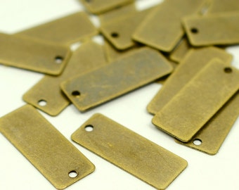May include: A collection of small, rectangular metal tags with a distressed, antique brass finish. Each tag has a single hole at one end. The tags are scattered on a white surface, creating a textured, close-up view.