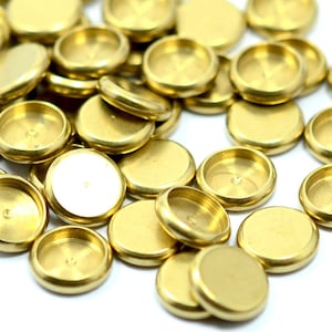 Puede incluir: Una colección de pequeños componentes metálicos redondos de color latón. Cada pieza tiene una parte superior plana y una hendidura circular poco profunda. Los componentes están dispersos sobre una superficie blanca, creando una disposición visualmente atractiva.