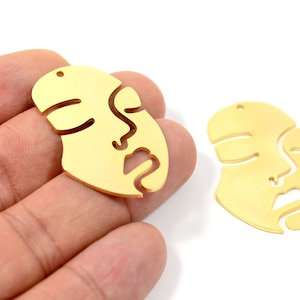 May include: Two gold-colored metal face-shaped charms with a cutout design. The charms have a hole at the top for attaching to jewelry.
