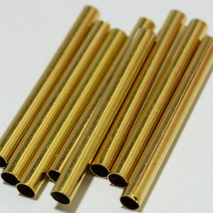 May include: A group of ten gold-colored metal tubes, all of the same size and shape, are arranged in a pile on a white surface.
