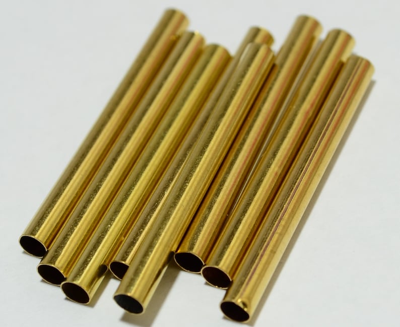 May include: A group of ten gold-colored metal tubes, all of the same size and shape, are arranged in a pile on a white surface.