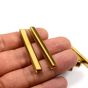 Puede incluir: Cuatro terminales de correa de metal dorado con un borde decorativo. Los terminales de correa est&aacute;n en una mano.