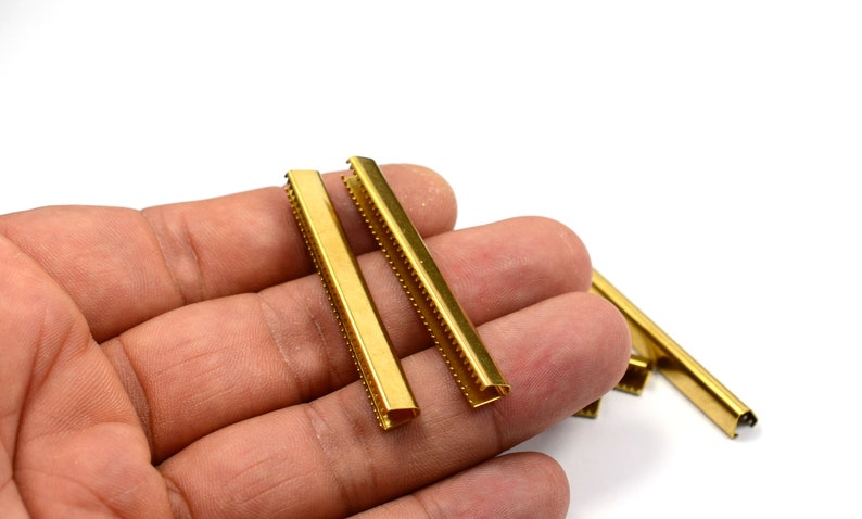 Puede incluir: Cuatro terminales de correa de metal dorado con un borde decorativo. Los terminales de correa est&aacute;n en una mano.