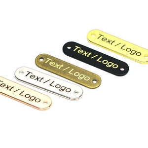 K&ouml;nnte beinhalten: F&uuml;nf Metall-Etiketten in verschiedenen Farben, darunter Gold, Silber, Bronze und Schwarz. Jedes Etikett ist oval geformt mit abgerundeten Kanten und zwei L&ouml;chern. Der Text "Text/Logo" ist auf jedem Etikett graviert.