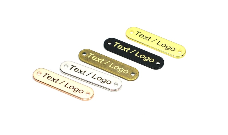K&ouml;nnte beinhalten: F&uuml;nf Metall-Etiketten in verschiedenen Farben, darunter Gold, Silber, Bronze und Schwarz. Jedes Etikett ist oval geformt mit abgerundeten Kanten und zwei L&ouml;chern. Der Text "Text/Logo" ist auf jedem Etikett graviert.