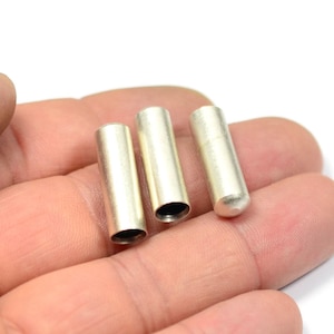 May include: Three silver-colored, cylindrical metal tubes with rounded ends. The tubes are held in a hand against a white background. The tubes are open at one end, revealing a dark interior.