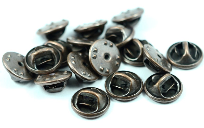 Puede incluir: Un mont&oacute;n de 15 respaldos de alfiler de metal de color bronce antiguo. Los respaldos de alfiler tienen una base redonda y plana con un anillo elevado alrededor del borde. Los respaldos de alfiler est&aacute;n dise&ntilde;ados para ser utilizados con una variedad de alfileres y broches.