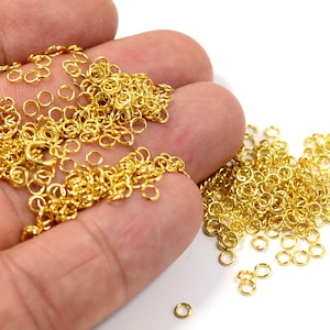 May include: A large pile of gold jump rings, a type of jewellery finding, are shown in a hand. The jump rings are small and round, and they are perfect for connecting different pieces of jewellery together.