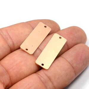May include: Two gold-colored metal rectangular tags with two holes each. The tags are on a hand.