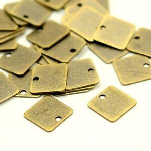 May include: A pile of antique brass square charms with a hole in each one. The charms are scattered on a white surface.
