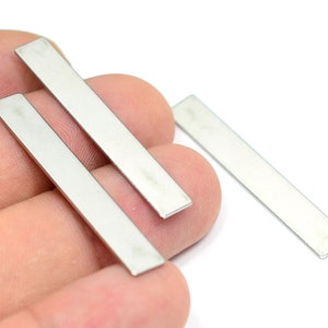 May include: Three silver metal bars, approximately 2 inches long, are shown on a hand. The bars are smooth and rectangular.